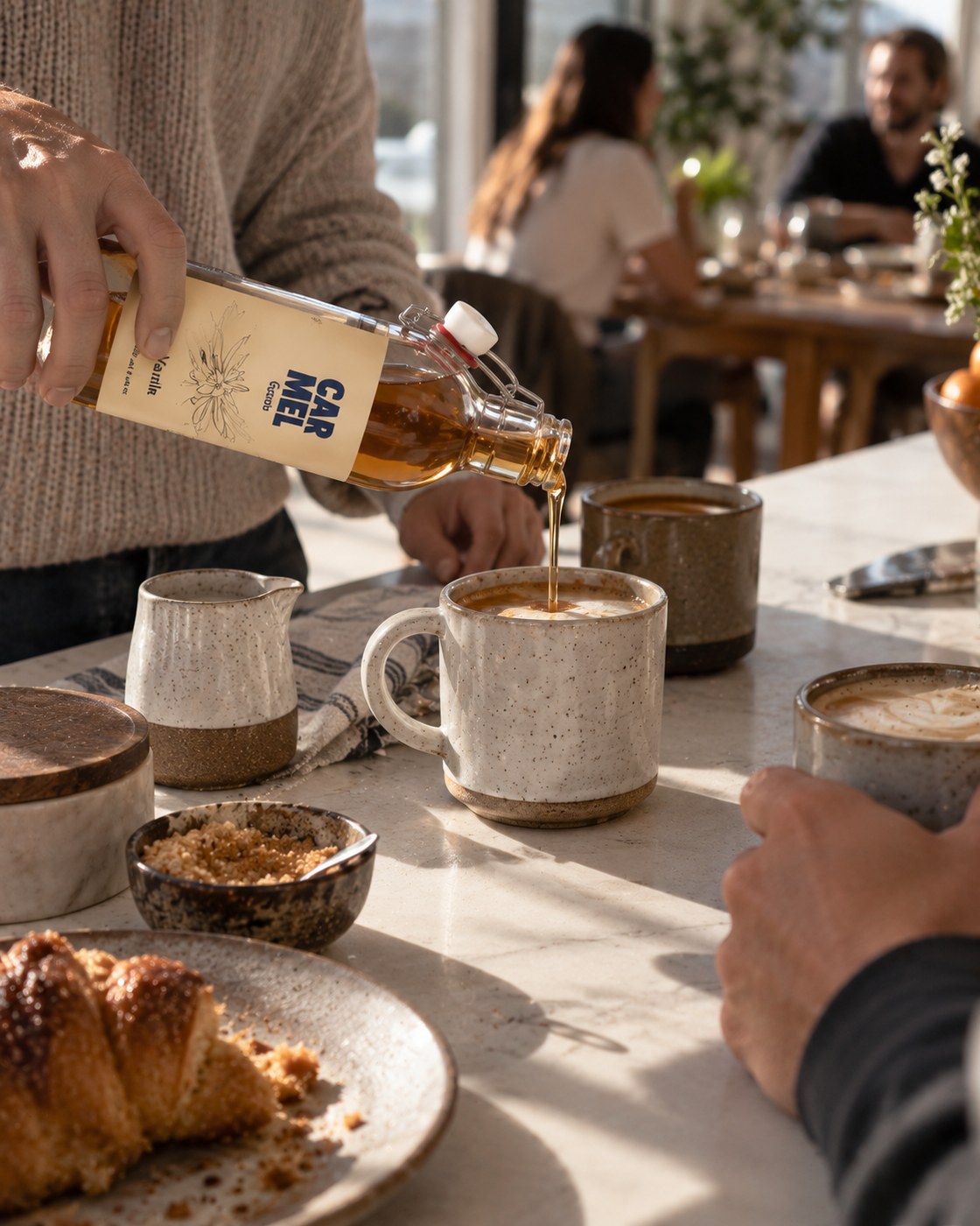 Friends gathered at a sunlit table while CARMEL Goods Vanilla Syrup is poured into a latte